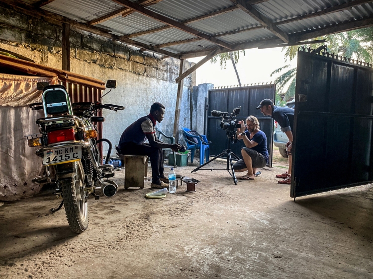 Photographer, Jason Florio, films an interview with a young Gambian man who is sitting in by an open gate, making attaya tea, next to a motorcycle, The Gambia, West Africa , Image ©Helen Jones-Florio