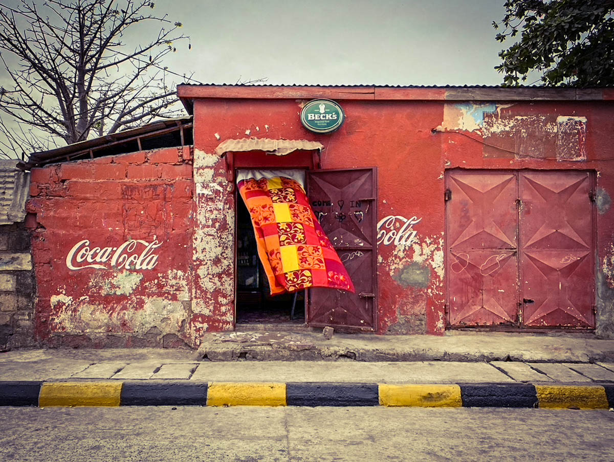 #GambiaDoors - a brightly coloured curtain blows in the doorway of a local bar, a Becks beer sign above the door, painted red with Coca Cola written on the wall, Banjul, The Gambia ©Helen Jones-Florio