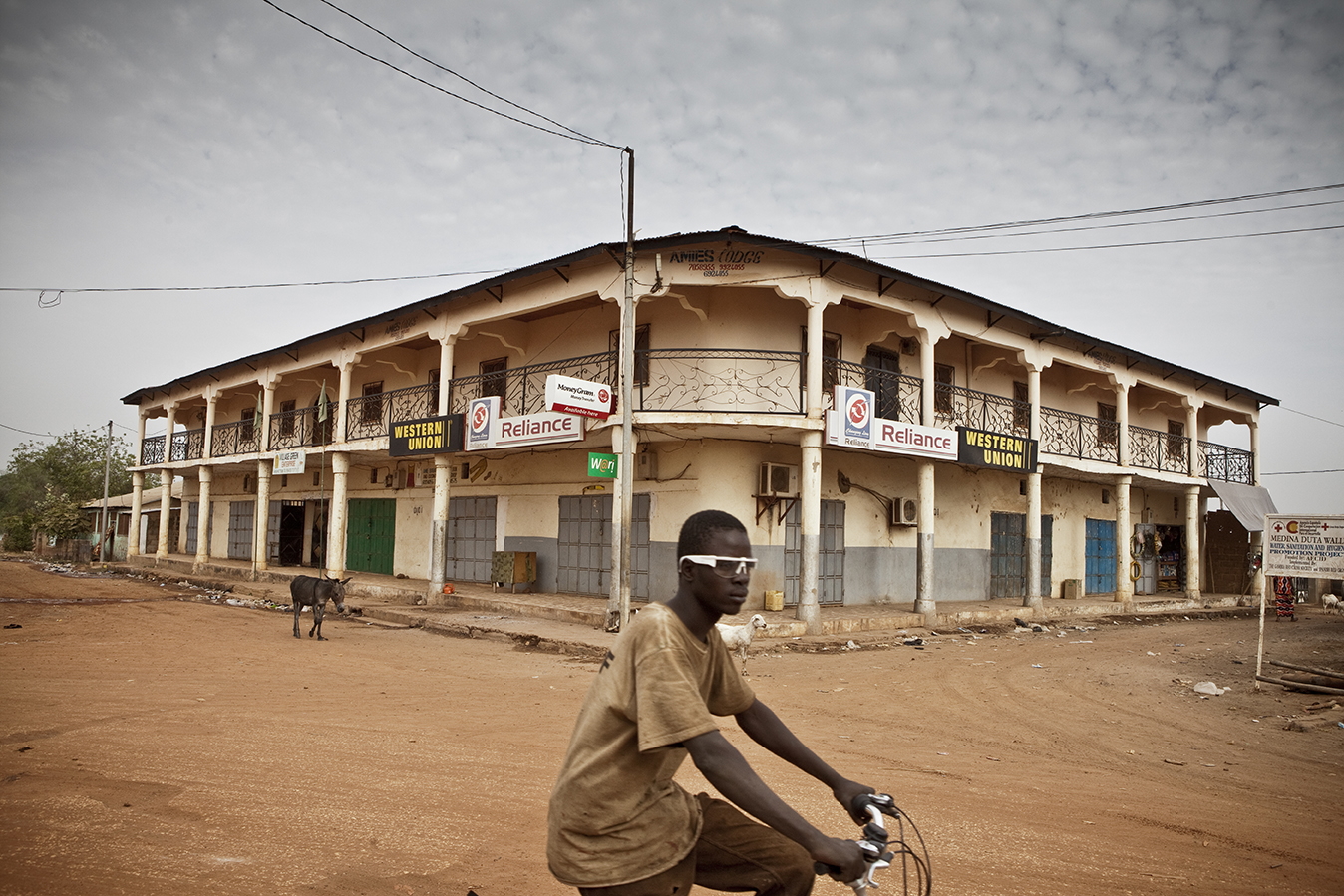 A boy in white framed sunglasses riding a bicycle past an old colonial style building in The Gambia, West Africa © Jason Florio